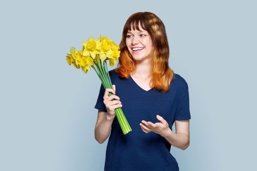 Young teenage red-haired female with bouquet of yellow flowers on light gray color background. Spring, beauty, holidays, youth concept