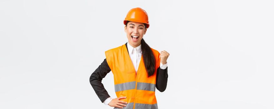 Successful businesswoman, construction manager in safety helmet, fist pump and say yes. Female asian architect celebrating great news, achieve goal at building zone, white background.