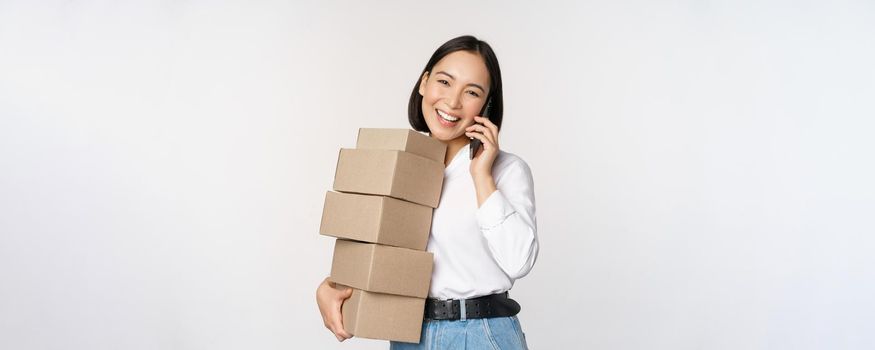 Image of young asian businesswoman answer phone call while carrying boxes for delivery, posing against white background.