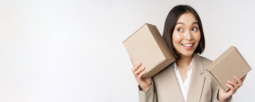 Image of asian saleswoman shaking boxes and guessing whats inside, smiling thoughtful, standing over white background. Copy space