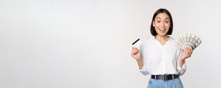 Portrait of enthusiastic asian woman holding money in cash and credit card, smiling amazed at camera, white background.