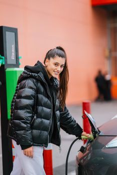 Young beautiful woman traveling by electric car having stop at charging station standing plugging cable browsing internet on smartphone smiling joyful while charing