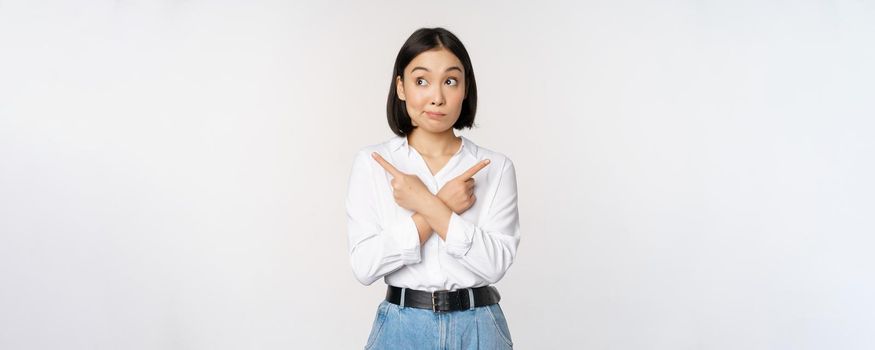 Indecisive asian woman pointing fingers sideways, pointing fingers and looking clueless, confused with choices, standing over white background.