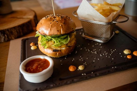 Closeup of fresh burger with basket of French fries on wooden table with bowl of tomato sauce