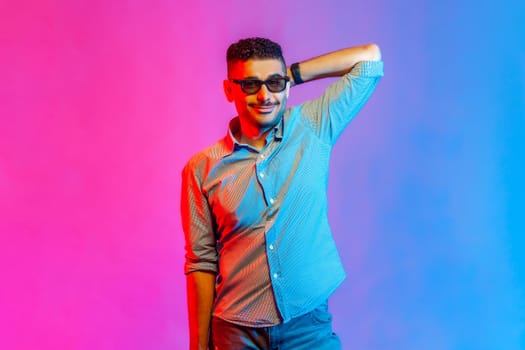 Portrait of man in shirt and glasses standing with raised hand and looking at camera with positive smile and optimistic expression. Indoor studio shot isolated on colorful neon light background.