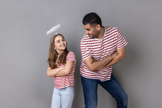 Portrait of smiling joyful father and daughter in striped T-shirts standing with folded hands and looking at each other, family expressing happiness. Indoor studio shot isolated on gray background.