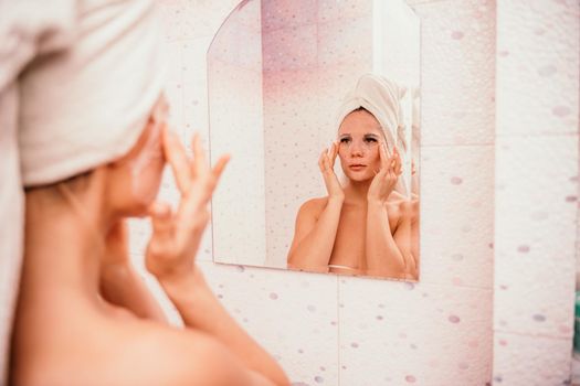Young beautiful woman using a moisturizing facial mask after taking a bath. Pretty attractive girl in a towel on her head stands in front of a mirror in a home bathroom. Daily hygiene and skin care.