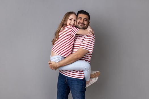 Portrait of father and daughter in striped T-shirts holding his child, feeling love and deep devotion, warm relationships, love care, fathers day. Indoor studio shot isolated on gray background.