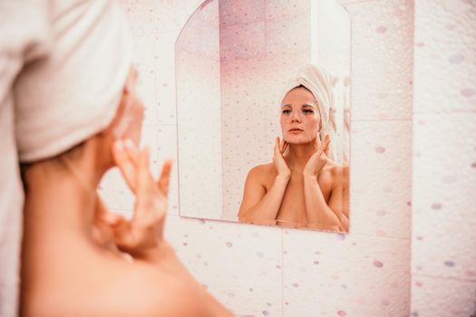 Young beautiful woman using a moisturizing facial mask after taking a bath. Pretty attractive girl in a towel on her head stands in front of a mirror in a home bathroom. Daily hygiene and skin care.