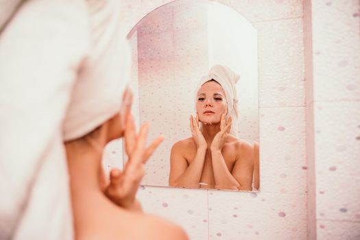 Young beautiful woman using a moisturizing facial mask after taking a bath. Pretty attractive girl in a towel on her head stands in front of a mirror in a home bathroom. Daily hygiene and skin care.