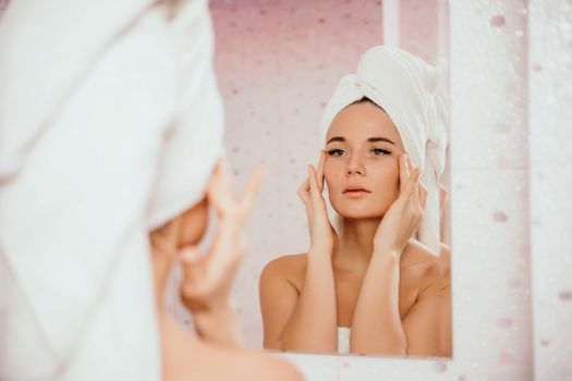 Young beautiful woman using face cream moisturizing lotion after bath. Pretty attractive girl in a towel on her head stands in front of a mirror in a home bathroom. Daily hygiene and skin care.