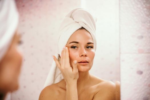 Young beautiful woman using face cream moisturizing lotion after bath. Pretty attractive girl in a towel on her head stands in front of a mirror in a home bathroom. Daily hygiene and skin care.