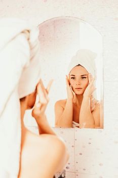 Young beautiful woman using face cream moisturizing lotion after bath. Pretty attractive girl in a towel on her head stands in front of a mirror in a home bathroom. Daily hygiene and skin care.