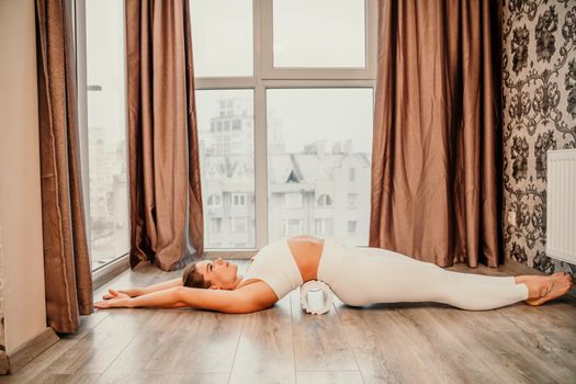 Adult athletic woman, in white bodysuit, performing fascia exercises on the floor - caucasian woman using a massage foam roller - a tool to relieve tension in the back and relieve muscle pain - the concept of physiotherapy and stretching training.