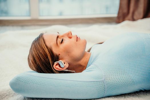 Side view portrait of relaxed woman listening to music with headphones lying on carpet at home. She is dressed in a blue tracksuit