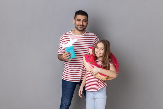 Portrait of joyful happy father and daughter in striped T-shirts are going to have vocation, holding rubber ring, passport and paper plane. Indoor studio shot isolated on gray background.