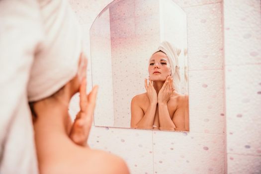 Young beautiful woman using a moisturizing facial mask after taking a bath. Pretty attractive girl in a towel on her head stands in front of a mirror in a home bathroom. Daily hygiene and skin care.