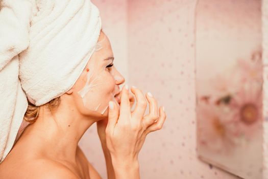 Young beautiful woman using a moisturizing facial mask after taking a bath. Pretty attractive girl in a towel on her head stands in front of a mirror in a home bathroom. Daily hygiene and skin care.