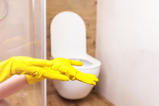 Close-up of two female hands in protective yellow rubber cleaning gloves. The background is a beautiful, modern bathroom and toilet in a minimalist style. Copy paste for your object, product
