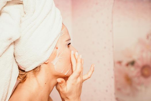 Young beautiful woman using a moisturizing facial mask after taking a bath. Pretty attractive girl in a towel on her head stands in front of a mirror in a home bathroom. Daily hygiene and skin care.