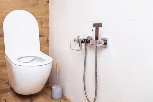 Fashionable bathroom in a minimalist style interior. Close-up of a white toilet bowl and a chrome-plated hygienic shower on the wall. The concept of hygiene, body care. Modern sanitary equipment
