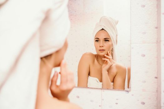 Portrait of young girl with towel on head in white bathroom looks and touches her face in the mirror and enjoys youth and hydration. Natural beauty, home care for problem skin.