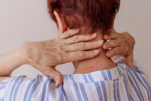 close-up, on a white background. An elderly white woman in a blue T-shirt is massaging her neck. the woman stands with her back, without personality. Neurological diseases of the spine, tremor