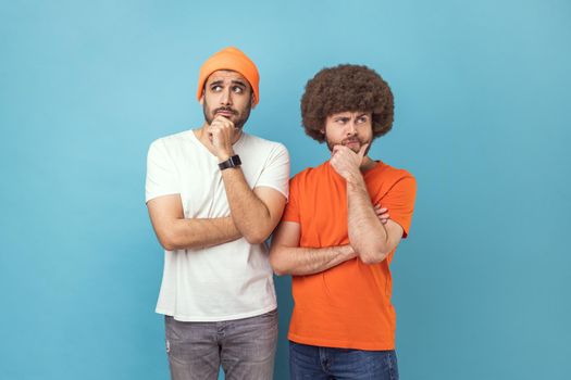 Portrait of two young adult hipster men holding chin with hands, need to think, planning strategy, thinking over ideas for startup. Indoor studio shot isolated on blue background.