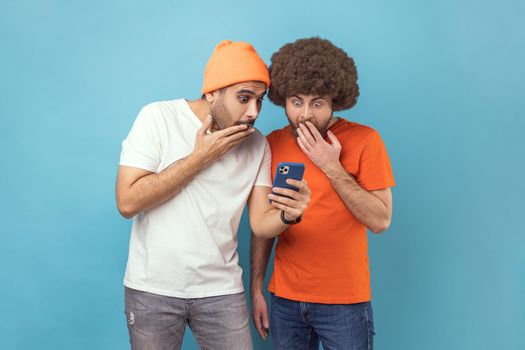 Portrait of two attractive shocked young adult hipster men standing, using smartphone, reading breaking news in social networks, covering open mouth. Indoor studio shot isolated on blue background.
