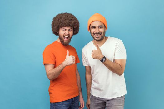 Well done, excellent job. Two young adult men showing thumbs up, gesturing like to camera and smiling broadly, expressing approval, positive feedback. Indoor studio shot isolated on blue background.