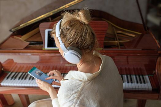 young caucasian blonde girl in headphones plays the vintage piano at home. A woman watches a music lesson on her smartphone. Concept of online learning and new hobby