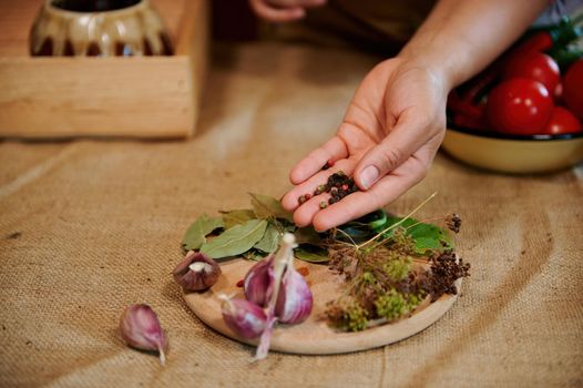 Details: female hand holds peppercorns above wooden chopping board with garlic cloves, bay and fresh dill leaves. Fragrant culinary herbs and ingredients on kitchen table with rustic linen tablecloth