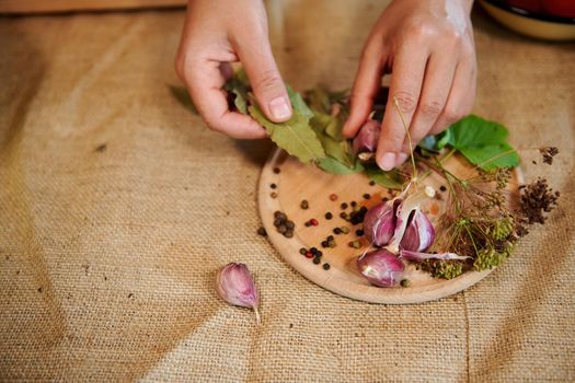 Still life. Housewife's hands, holding fragrant culinary herbs above a chopping board with laid out garlic cloves, fresh dill leaves, dry bay leaves and scattered peppercorns on a linen tablecloth