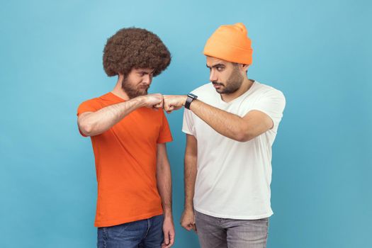 Portrait of two serious young adult hipster men standing and looking at each other with aggressive look and with bump gesture, fighting. Indoor studio shot isolated on blue background.