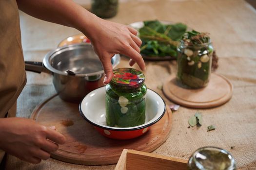 Close-up of a housewife's hand, putting a sterile lid on a glass jar of freshly pickled organic cucumbers. Homemade canned food. Preparing marinated delicacy for the winter season. Pickling.