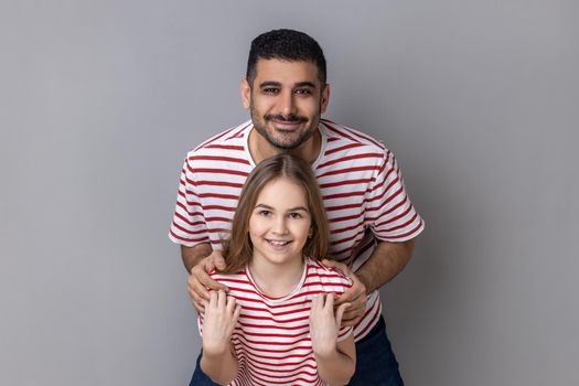 Portrait of delighted smiling father and daughter in striped T-shirts standing and looking at camera, dad embracing little kid, expressing happiness. Indoor studio shot isolated on gray background.