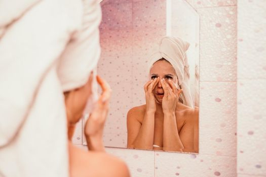 Young beautiful woman using a moisturizing facial mask after taking a bath. Pretty attractive girl in a towel on her head stands in front of a mirror in a home bathroom. Daily hygiene and skin care.