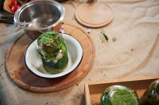 Top view of a jar with fresh organic cucumbers and fragrant culinary herbs in an enamel bowl and a saucepan with boiling marinade on a kitchen table with linen tablecloth. Still life with canned food