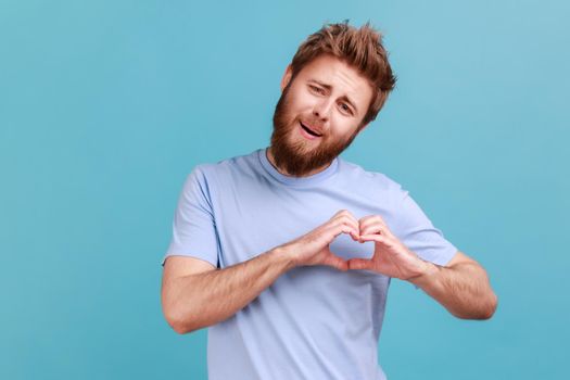 Portrait of handsome bearded man holding hands in shape of heart showing romantic gesture, love confession, valentines day celebration. Indoor studio shot isolated on blue background.