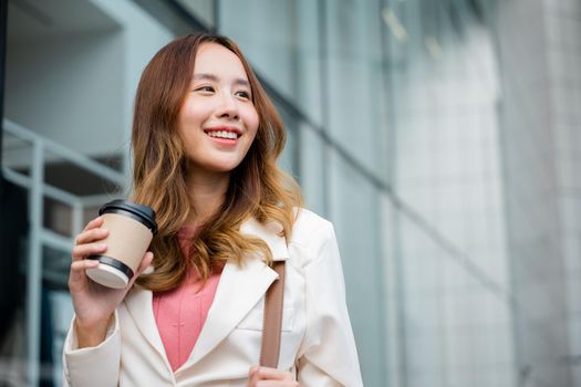 Happy business woman smiling hold paper cup of hot drink outdoor walking on street, Asian businesswoman holding coffee cup takeaway going to work she walking near her office building outside