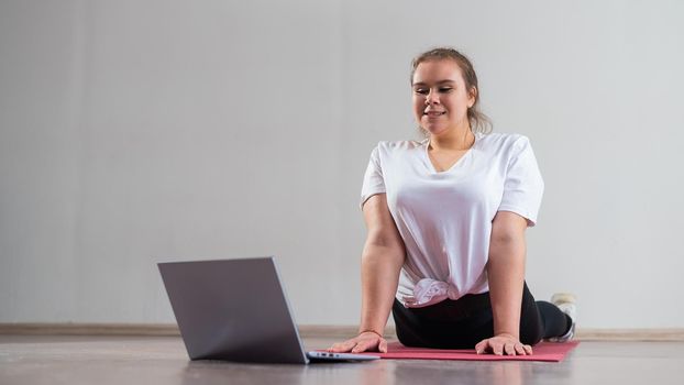 Young Plus Size Woman Stretching At Home Online. Flexible girl practices yoga and watches an online course on a laptop.