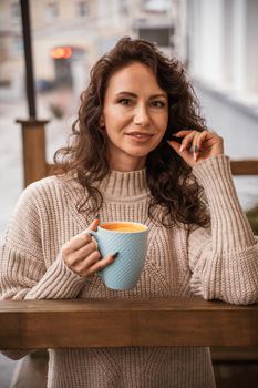 A middle-aged woman in a beige sweater with a blue mug in her hands is in a street cafe on the veranda.