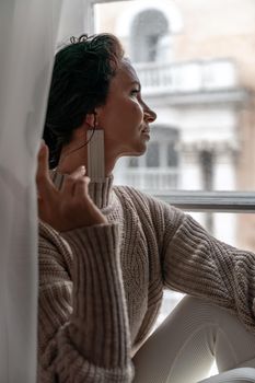 Free time for rest. Profile of a beautiful woman sitting on a white window sill at home, holding a cotton plant in her hands. She is wearing a sweater, leggings and white boots