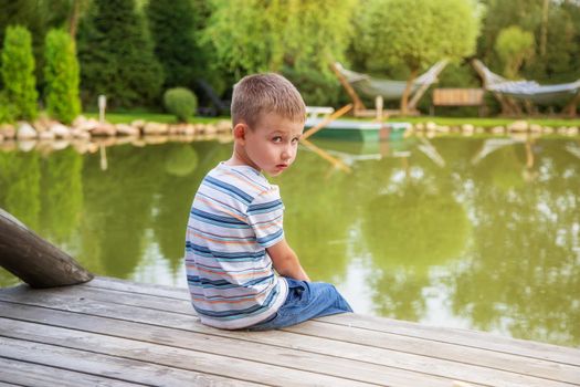 Little European boy in a striped T-shirt, sitting with his back. The child is sad alone. Sunny summer day. The concept of childhood, happiness and tranquility. June 1st,