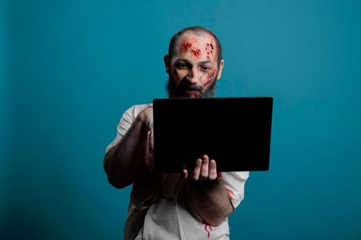 Dangerous halloween monster browsing internet on laptop in front of camera, holding computer and being scary creepy apocalyptic. Undead frightening evil zombie using pc with social media.