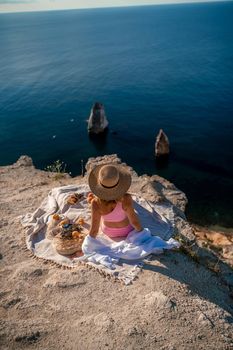 photo of a beautiful woman with long blond hair in a pink shirt and denim shorts and a hat having a picnic on a hill overlooking the sea.