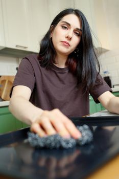 Tired young woman with a metal sponge for cleaning, washes a baking sheet in the kitchen, selective focus, vertical.