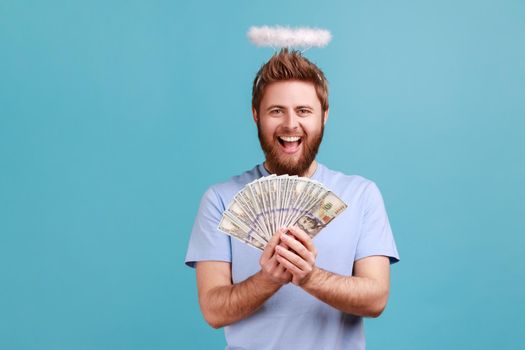 Portrait of excited bearded angelic man with holy nimbus over head holding fan of dollar bills and smiling happily, happy to have money. Indoor studio shot isolated on blue background.