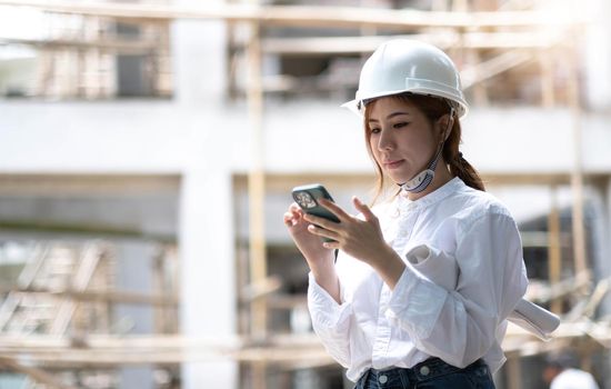 Architect with a blueprints at a construction site. Portrait of woman constructor wearing white helmet and safety yellow vest. Upset sad, skeptical, serious woman looking at the phone screen outdoors..