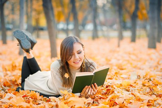 Mixed race woman lying on fall foliage and reading book in autumn park.